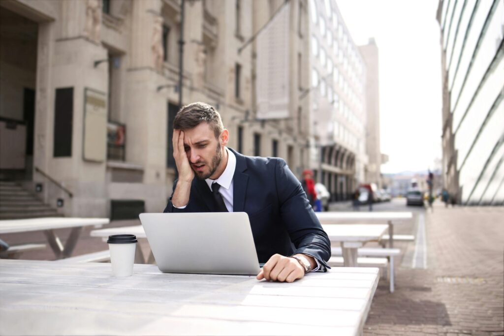 A businessman in a black suit looks stressed while working outdoors on a laptop with coffee.